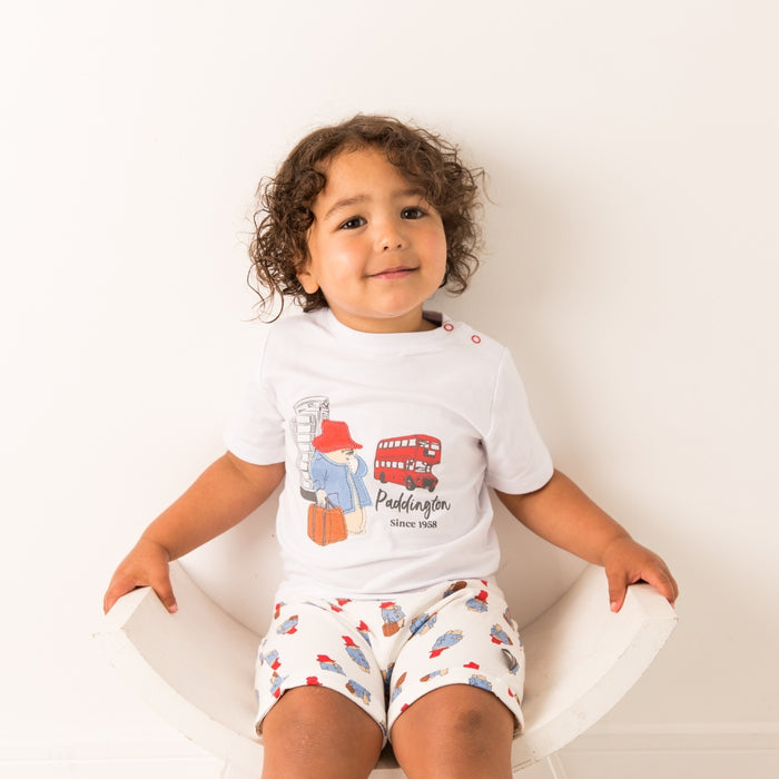 Child wearing a white t-shirt with Paddington Bear graphics and shorts sitting on a white chair.