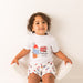 Child wearing a white t-shirt with Paddington Bear graphics and shorts sitting on a white chair.