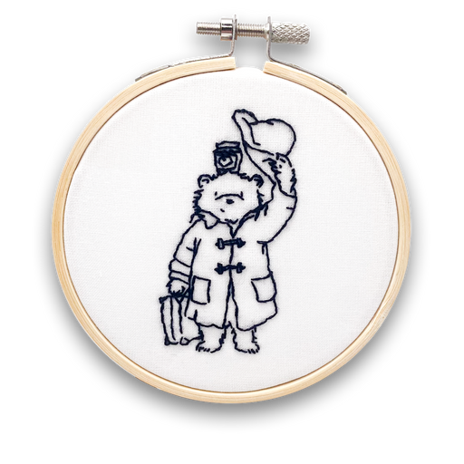 A mini embroidery kit depicting the character Paddington Bear holding a hat and a jar of marmalade, working on a piece of fabric.