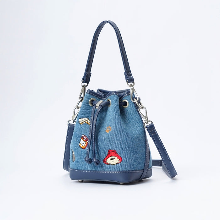 A blue denim bucket bag with a navy blue leather trim, featuring embroidered Paddington bear patches on the front, a top handle, and a detachable shoulder strap, set against a plain white background.