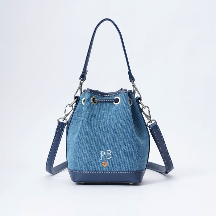 A blue denim bucket bag with navy blue leather trim, silver-tone hardware, and a white embroidered 'PB' monogram on the front, set against a plain white background.