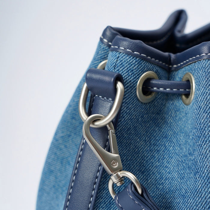 Close-up of a blue denim drawstring bucket bag with navy leather trim, silver-tone metal grommets, and a matching navy leather strap attached with a silver clasp.