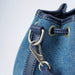 Close-up of a blue denim drawstring bucket bag with navy leather trim, silver-tone metal grommets, and a matching navy leather strap attached with a silver clasp.