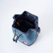 A blue denim bucket bag with navy leather trim, silver-tone metal eyelets, and embroidered decorative motifs including Paddington, shown from an overhead angle on a white background.