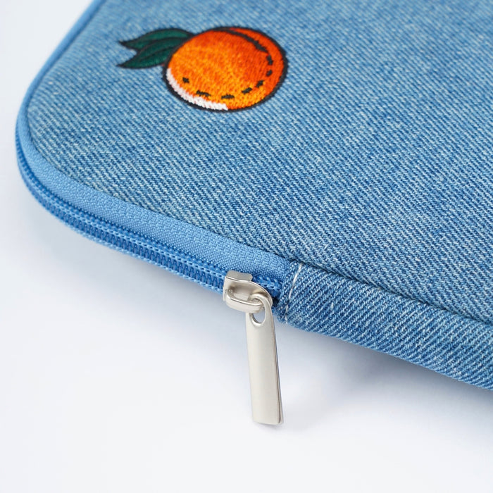 Close-up of a blue denim zippered laptop case featuring an embroidered orange with green leaves on the top corner and a silver metal zipper pull, set against a plain white background.