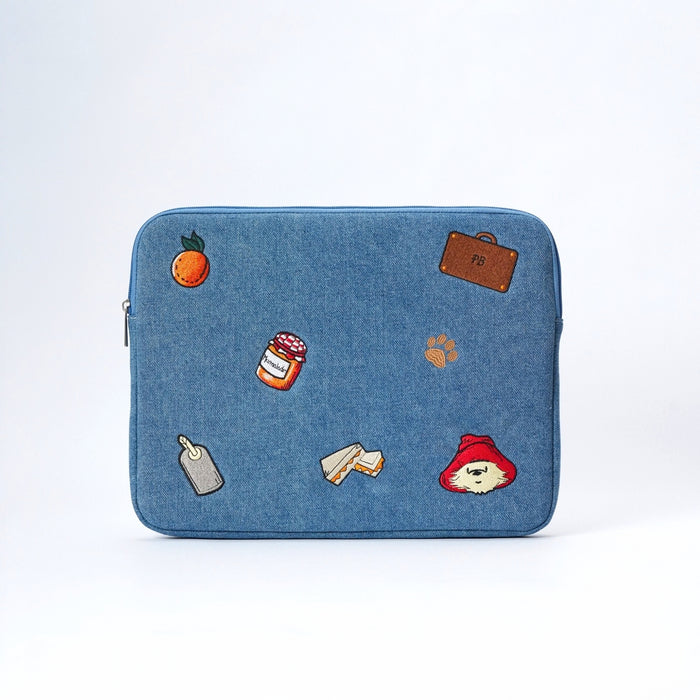 A blue denim laptop case featuring Paddington bear embroidery and props including an orange, suitcase, marmalade jar, pawprint, luggage tag, marmalade sandwich and Paddington's face.