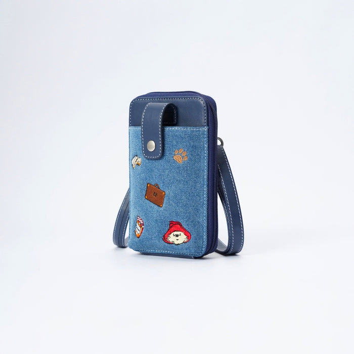 A blue denim phone pouch with dark navy blue trim, featuring a silver snap button closure and a wrist strap. The front pocket is decorated with several colorful embroidered patches, including a brown suitcase, a red hat, and a paw print.