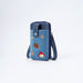 A blue denim phone pouch with dark navy blue trim, featuring a silver snap button closure and a wrist strap. The front pocket is decorated with several colorful embroidered patches, including a brown suitcase, a red hat, and a paw print.