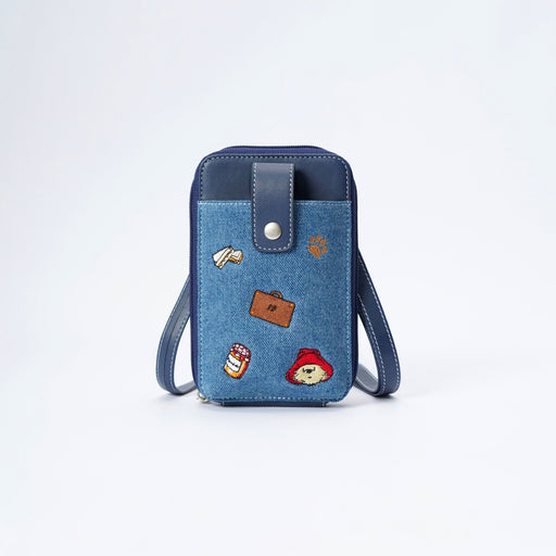 A blue denim phone bag with dark navy blue trim, featuring a silver snap button closure and a wrist strap. The front pocket is decorated with several colorful embroidered patches, including a brown suitcase, Paddington bear, and a paw print.