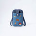 A blue denim phone bag with dark navy blue trim, featuring a silver snap button closure and a wrist strap. The front pocket is decorated with several colorful embroidered patches, including a brown suitcase, Paddington bear, and a paw print.