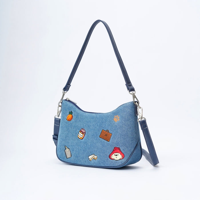 A blue denim shoulder bag with a dark blue leather strap and silver hardware, decorated with various colorful embroidered patches including an orange, a marmalade jar, and Paddington Bear.