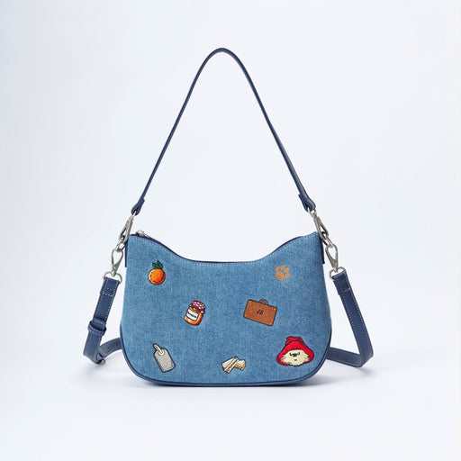 A blue denim crescent-shaped shoulder bag with a long strap and silver hardware, decorated with small colorful embroidered patches including an orange, a jar, a suitcase, and Paddington Bear.