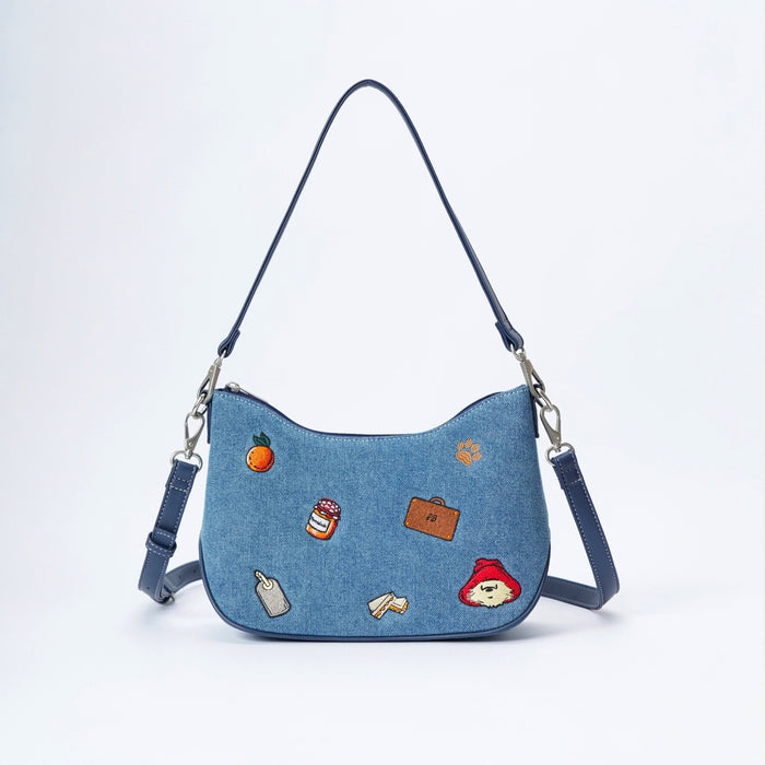 A blue denim crescent-shaped shoulder bag with a long strap and silver hardware, decorated with small colorful embroidered patches including an orange, a jar, a suitcase, and Paddington Bear.