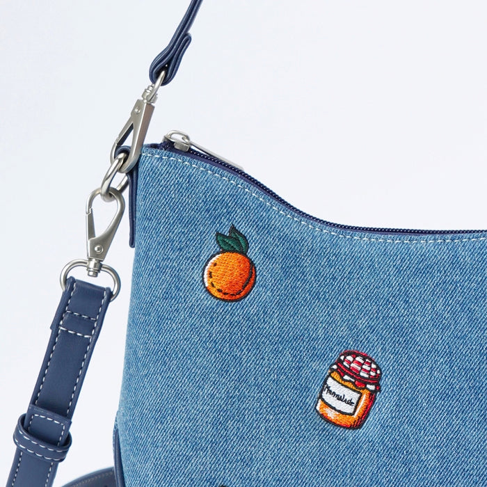 Close-up of a blue denim shoulder bag with embroidered details, including an orange with green leaves and a small marmalade jar with a red and white lid and label.
