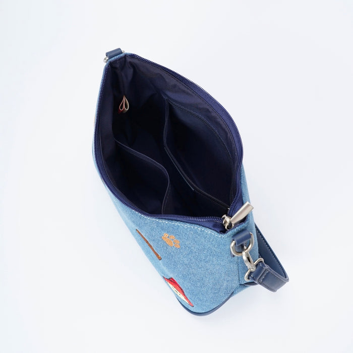 A light blue denim shoulder bag shown open from a top-down angle, revealing a dark navy interior, silver-tone hardware, and a small red embroidered Paddington Bear detail on the front.