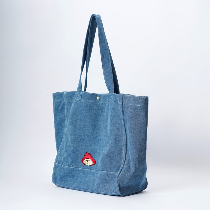 A large blue denim tote bag with dual shoulder straps and a small embroidered Paddington bear patch on the front side, photographed against a clean white studio background.