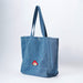 A large blue denim tote bag with dual shoulder straps and a small embroidered Paddington bear patch on the front side, photographed against a clean white studio background.