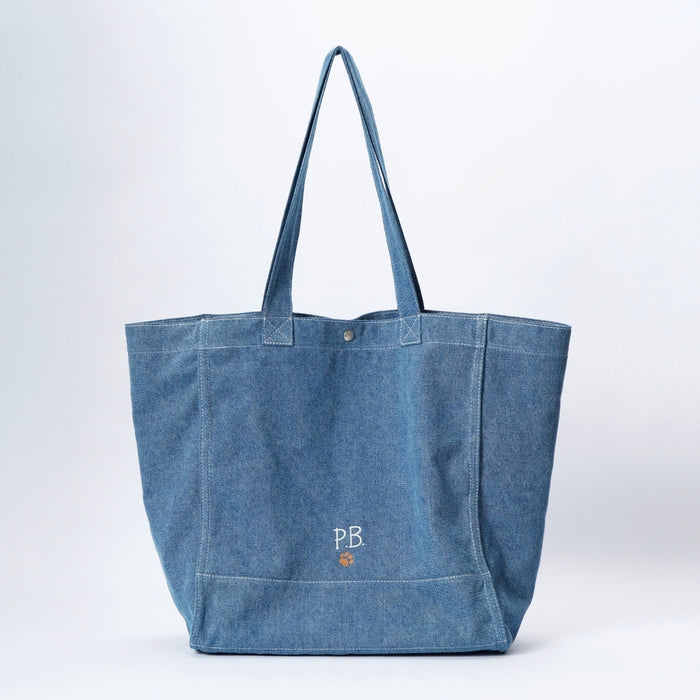 An oversized blue denim tote bag with dual shoulder straps, featuring a small white embroidered 'PB' logo and a tiny orange pawprint on the front pocket, set against a light gray studio background.