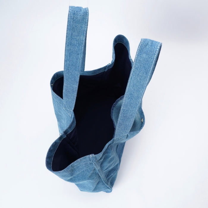 A blue denim tote bag with wide shoulder straps, shown from an overhead angle against a clean white background.