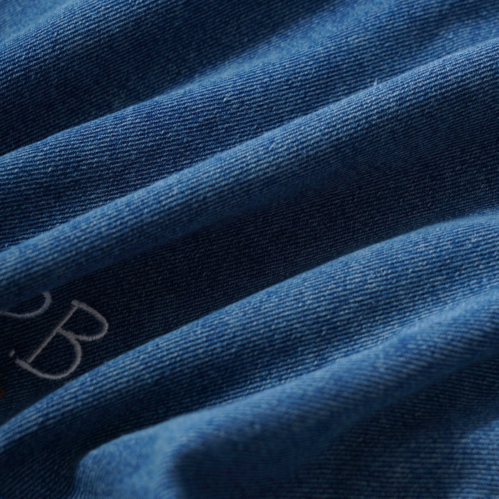 Close-up of blue denim fabric with soft folds and a small embroidered letter 'P.B.' visible on the left side.