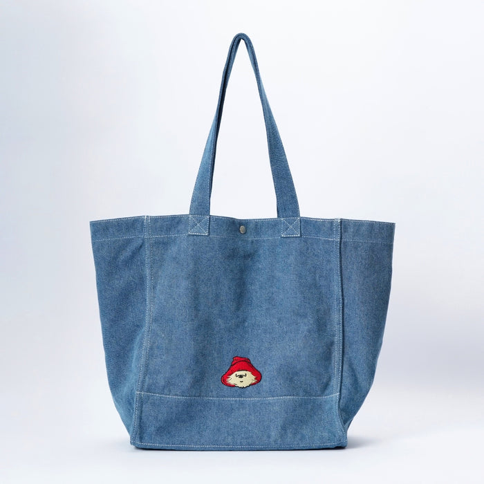 A large blue denim tote bag with long shoulder straps and a small embroidered Paddington bear patch on the front side, photographed against a clean white studio background.