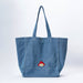 A large blue denim tote bag with long shoulder straps and a small embroidered Paddington bear patch on the front side, photographed against a clean white studio background.