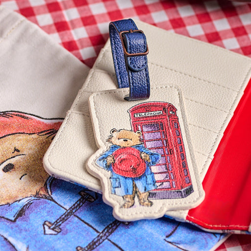 Paddington Bear Luggage Tag with blue leather strap and phone box artwork laying on top of a wallet