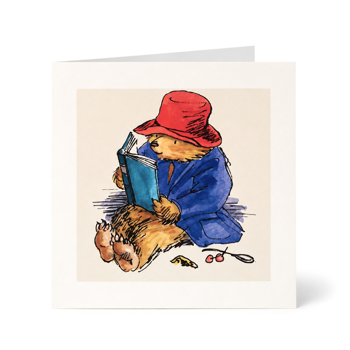 A square greeting card featuring a watercolor illustration of Paddington Bear reading a blue book, wearing his signature red hat and blue jacket, with cherries on the floor. The card is shown standing upright against a clean white background.