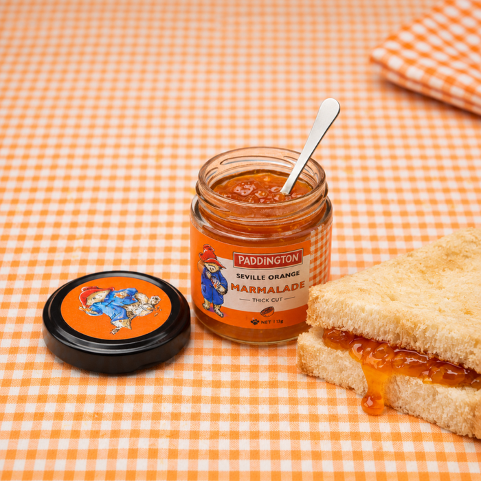 Jar of Paddington Seville Orange Marmalade with a spoon and a sandwich on an orange checkered tablecloth.
