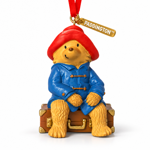 A Paddington bear ornament sitting on a brown suitcase, wearing a red hat and blue coat, with a gold tag reading PADDINGTON and a red hanging ribbon, on a white background.