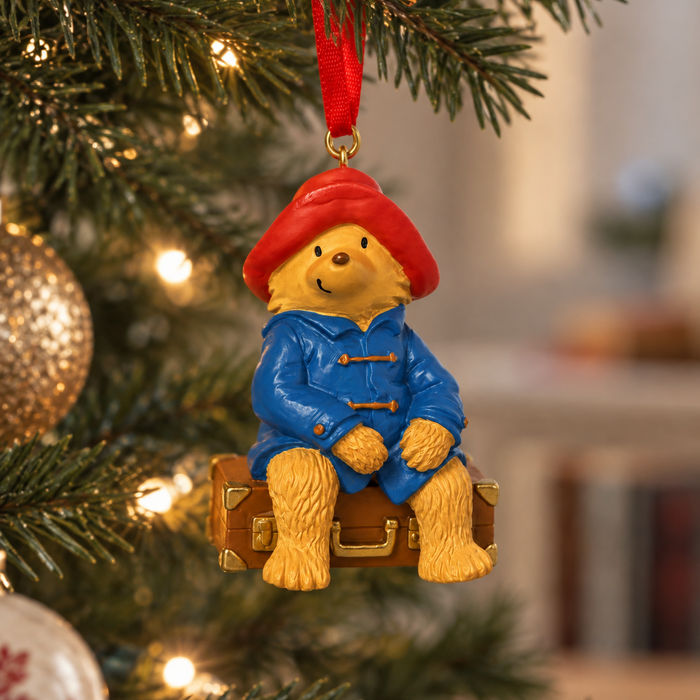 A Christmas ornament shaped like Paddington Bear sitting on a brown suitcase, hanging from a red ribbon on a lit Christmas tree. The background is a soft, blurred indoor setting with warm lights.