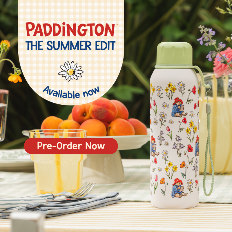 A white insulated water bottle with a light green lid and wrist strap, decorated with a colorful Paddington Bear floral print, displayed on a table next to a glass of orange juice. The background shows peaches on a platter and a blurred garden setting.