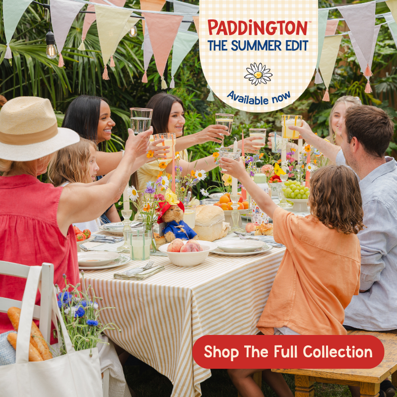 A group of people sitting around a decorated outdoor dining table, clinking glasses in a celebratory setting. The scene has colorful bunting, flowers, and food, with promotional text overlays reading 'PADDINGTON THE SUMMER EDIT Available now' and 'Shop The Full Collection'.