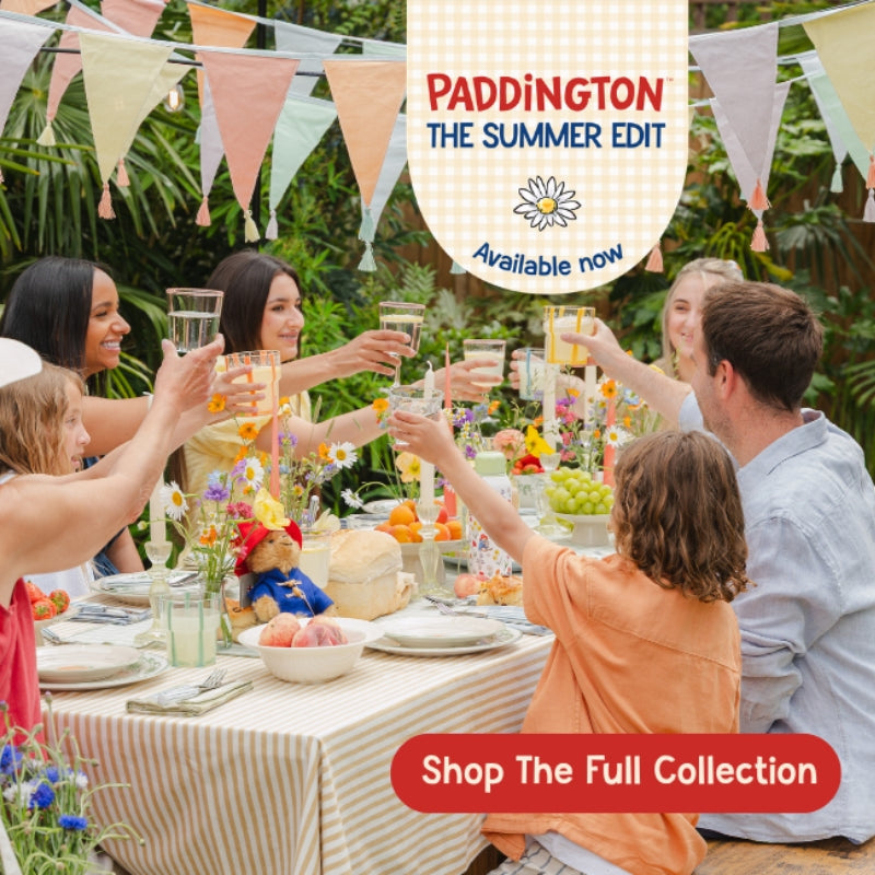 A family gathered around a table outdoors, raising glasses in a celebratory toast. The scene is decorated with pastel bunting flags, a striped tablecloth, and colorful floral arrangements.