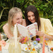 Two women reading a book together outdoors with flowers and drinks on a table.