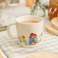 Ceramic mug with a Paddington Bear character writing and flowers on a checkered tablecloth.