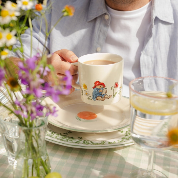 Person holding a mug with a cartoon Paddington bear design on a table with flowers and a glass.