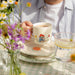 Person holding a mug with a cartoon Paddington bear design on a table with flowers and a glass.