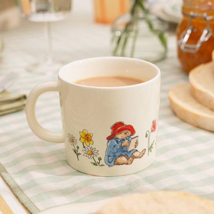 Ceramic mug with a Paddington Bear character writing and flowers on a checkered tablecloth.