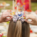 Person adjusting a colorful Paddington bear scrunchie on another person's hair with a blurred outdoor background