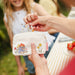 Small white pouch with Paddington Bear design held by a hand, with a blurred background of people and nature.