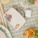 White pouch purse with Paddington Bear design on a checkered tablecloth with snacks and a glass of lemonade.