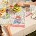 Table setting with a 'Paddington Bear' placemat, grapes, and glasses on a striped tablecloth.