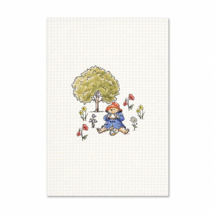 Illustration of paddington bear in a blue coat sitting under a tree with flowers and birds on a checkered background