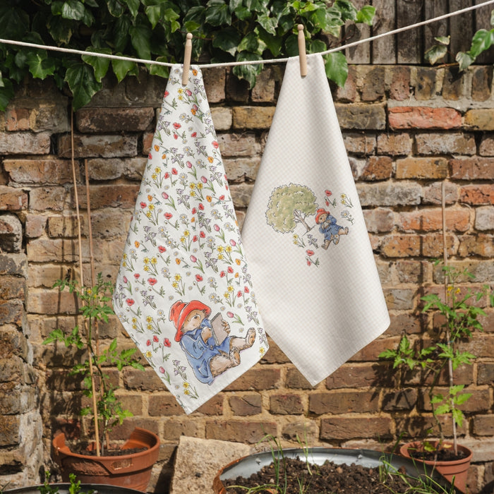 Two Paddington bear tea towels hanging on a line against a brick wall with plants.