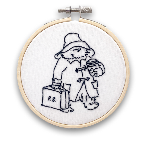 A mini embroidery kit depicting an illustration of Paddington bear with a suitcase and hat, holding a jar of marmalade, mounted on a hoop.