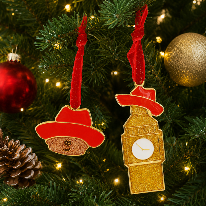 Two Paddington the musical Christmas tree ornaments hanging from a pine tree with warm lights: one shaped like Paddington bear wearing a red hat, and one shaped like Big Ben in gold with a red hat and white clock face. Red ribbons hang them from the branches. 