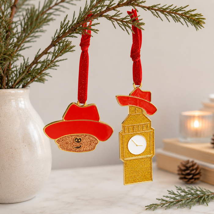 Two festive Christmas Paddington the Musical ornaments hanging from a pine branch: one shaped like Paddington bear in a red hat with a glittery brown face, and one shaped like Big Ben and a red hat above a gold tower clock with a white face. The scene is styled with a white vase, candles, books, and pinecones on a light surface. 