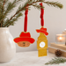 Two festive Christmas Paddington the Musical ornaments hanging from a pine branch: one shaped like Paddington bear in a red hat with a glittery brown face, and one shaped like Big Ben and a red hat above a gold tower clock with a white face. The scene is styled with a white vase, candles, books, and pinecones on a light surface. 