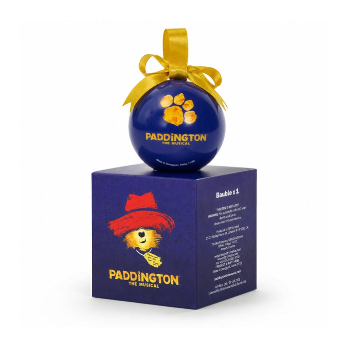 A blue Paddington The Musical Christmas bauble ornament resting on its matching blue packaging box, both featuring the yellow ribbon and character artwork, set against a plain white background.